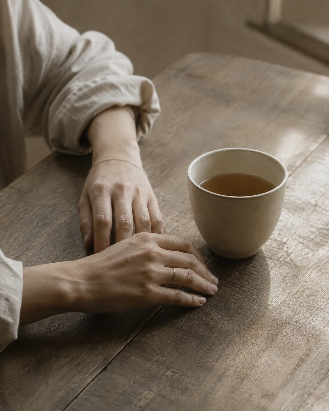 Two hands resting on a wooden table beside a cup of tea.